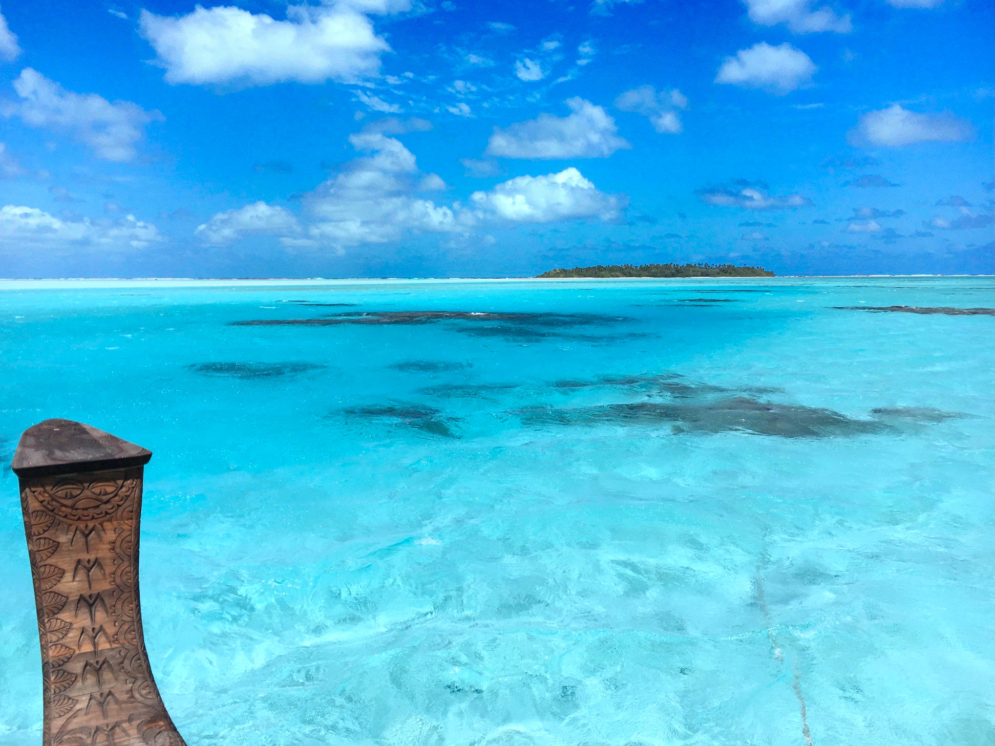 Das Bild zeigt die Aussicht von einem Boot auf das klare, blaue Wasser vor der Insel Aitutaki. Im Hintergrund ist eine Insel mit einem Palmenwald zu sehen.