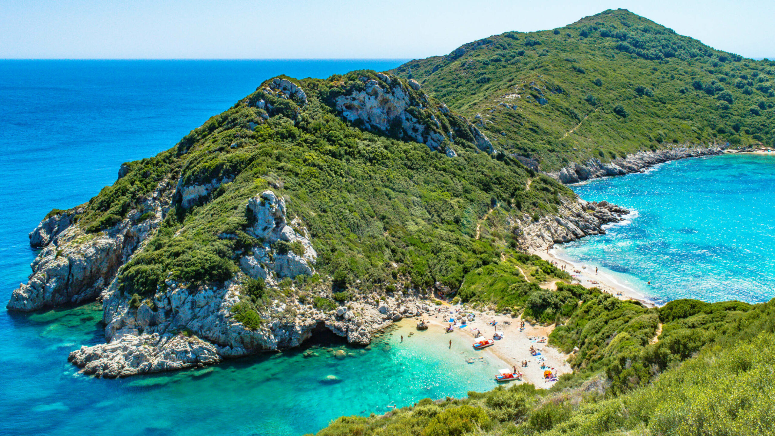 Das Bild zeigt den Zwillingsstrand auf Korfu. Mittig im Bild ist eine grüne Bergkette. An einem verengtem Abschnitt befinden sich links und rechts neben der Grünfläche Strände mit Menschen und Booten.