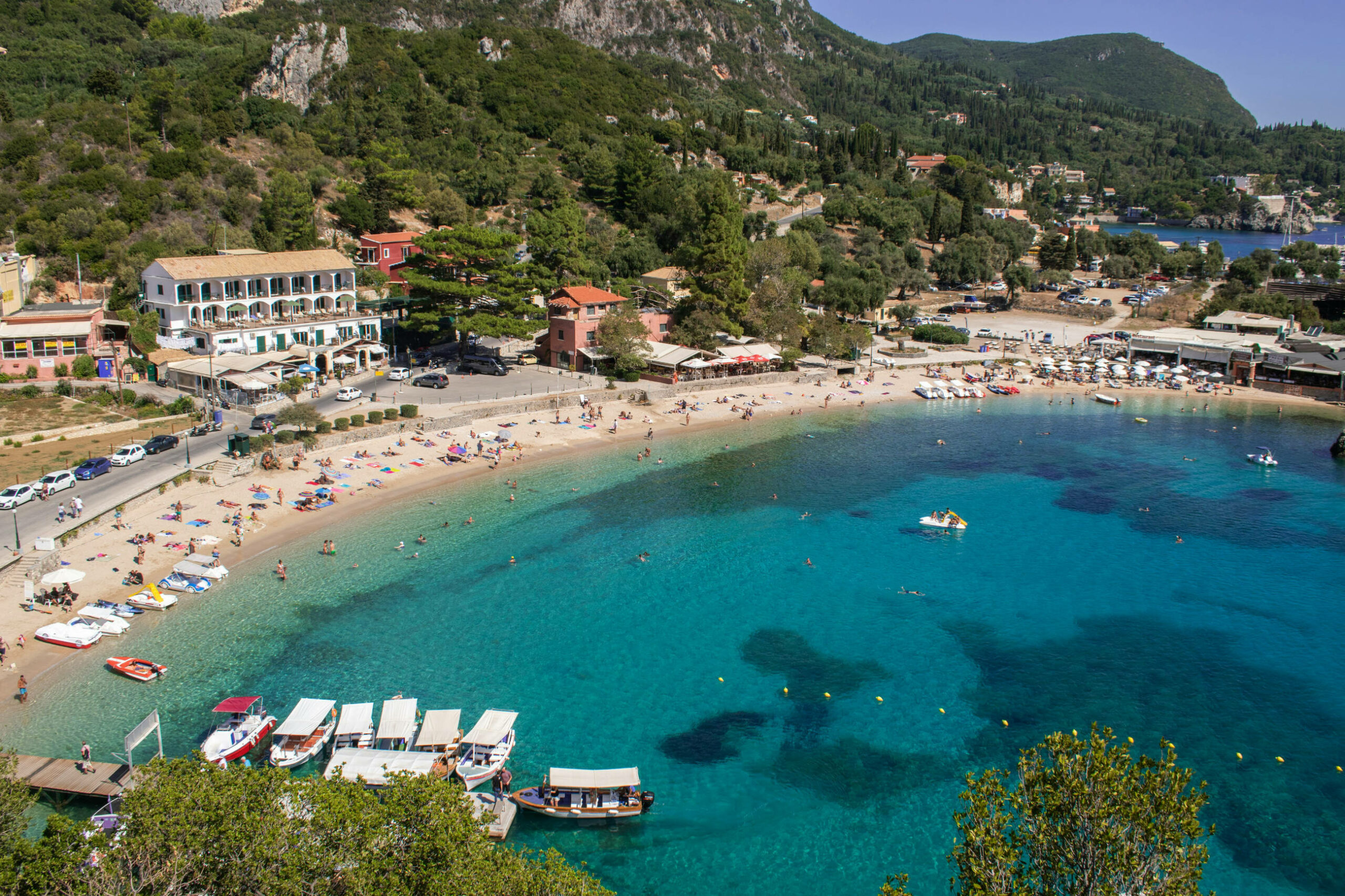Das Bild zeigt den Strand von Paleokastritsa auf Korfu. Im Vordergrund ist das Meer mit einigen Booten, Schwimmern und Begreunzungsbojen. Weiter hinten im Bild sind Menschen am Strand sowie Straßen mit Autos und Häuser. Im Hintergrund sind grüne Berge.