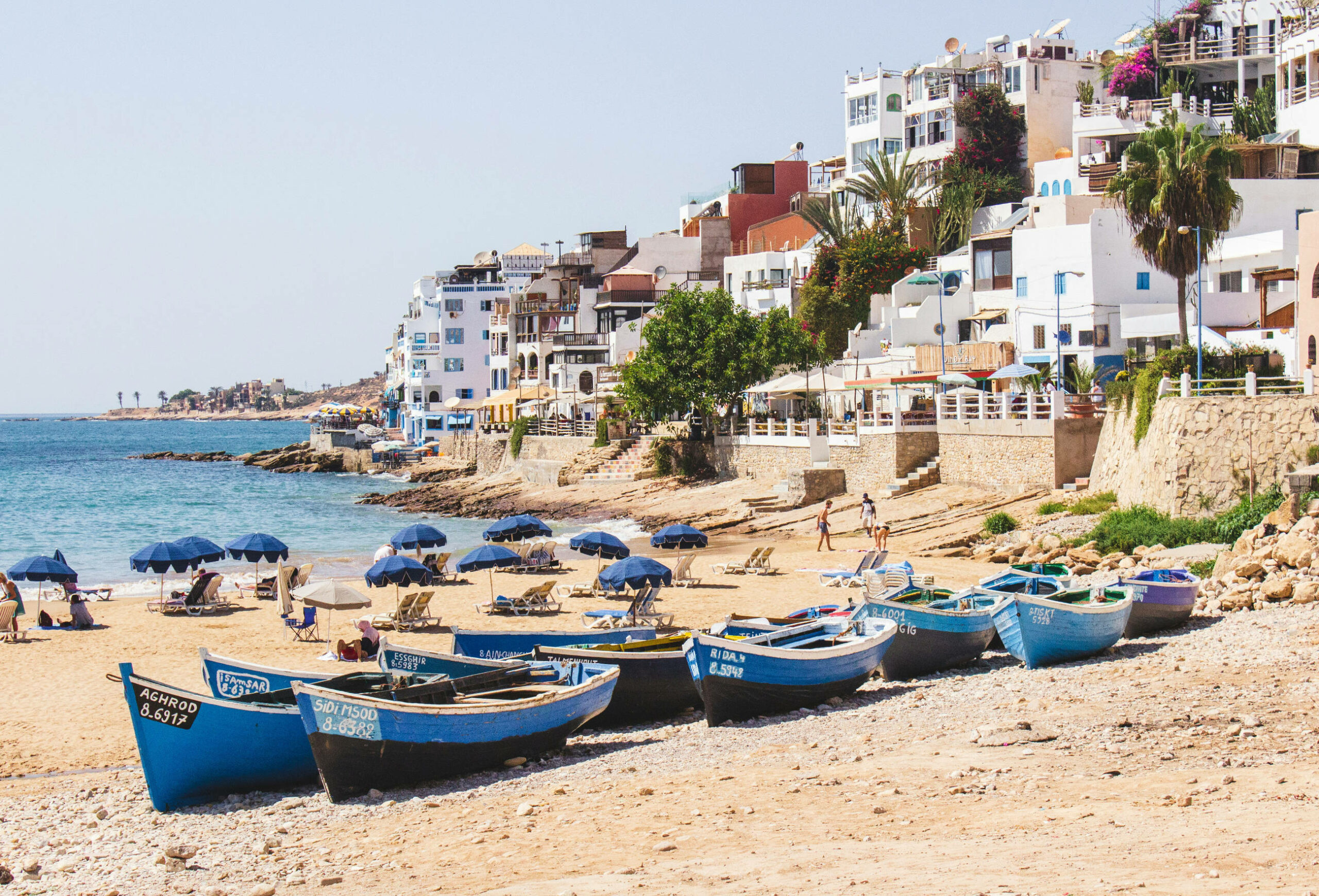 Das Bild zeigt einen Abschnitt von Taghazout Beach in Marokko. Im Vordergrund stehen einige blaue Boote. Im Hintergrund ist der Strand mit Liegen und Sonnenschirmen. Dazwischen laufen einige Menschen. Rechts im Bild sieht man viele weiße Häuser mit Pflanzen dazwischen.