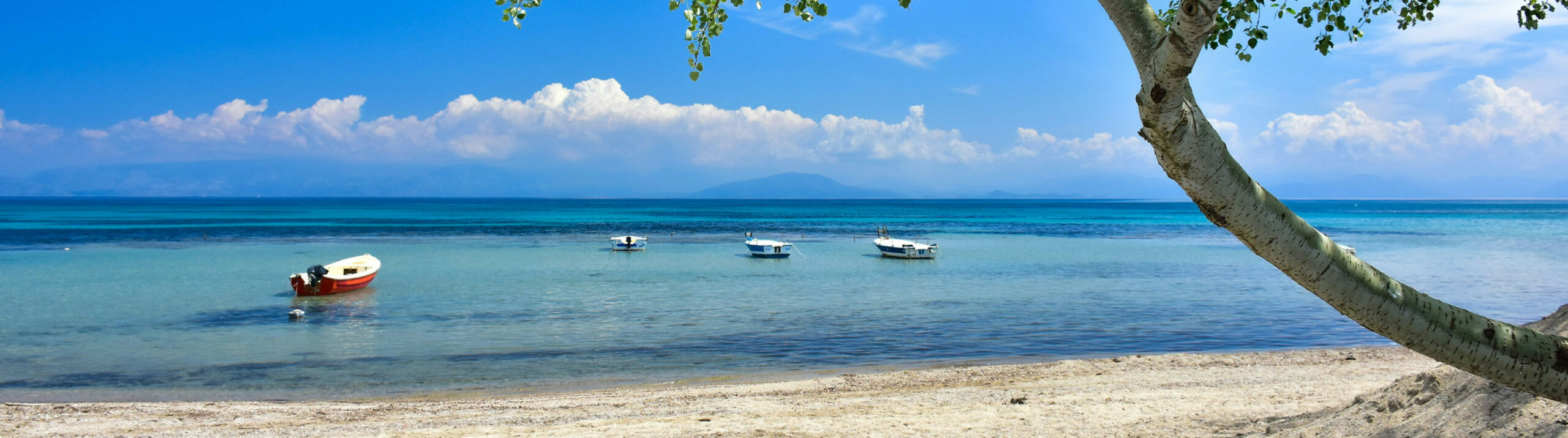 Das Bild zeigt einen Strand auf Korfu. Im Vordergrund sind Sand, Wasser und vier Boote zu sehen. Im Hintergrund sind Wolken am Himmel. In der weiten Ferne ist ein Berg zu sehen.