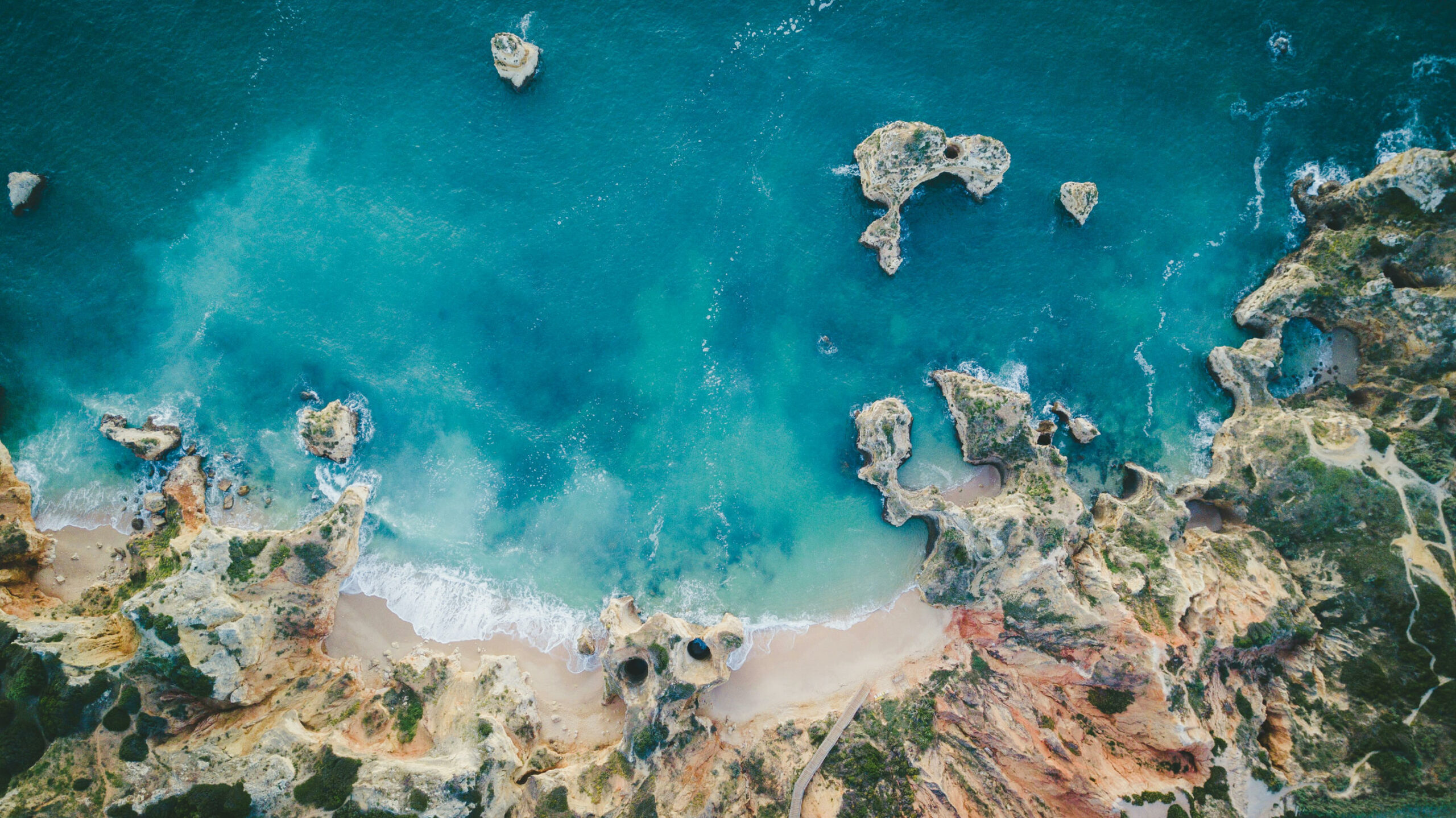Auf dem Bild sieht man einen Küstenabschnitt an der Algarve aus der Drohnenperspektive. Im mittleren bis oberen Teil des Bildes ist blaues Meer mit einigen großen Felsen. Im unteren Teil des Bildes sind drei Strandabschnitte und viele Felsformationen zu sehen.