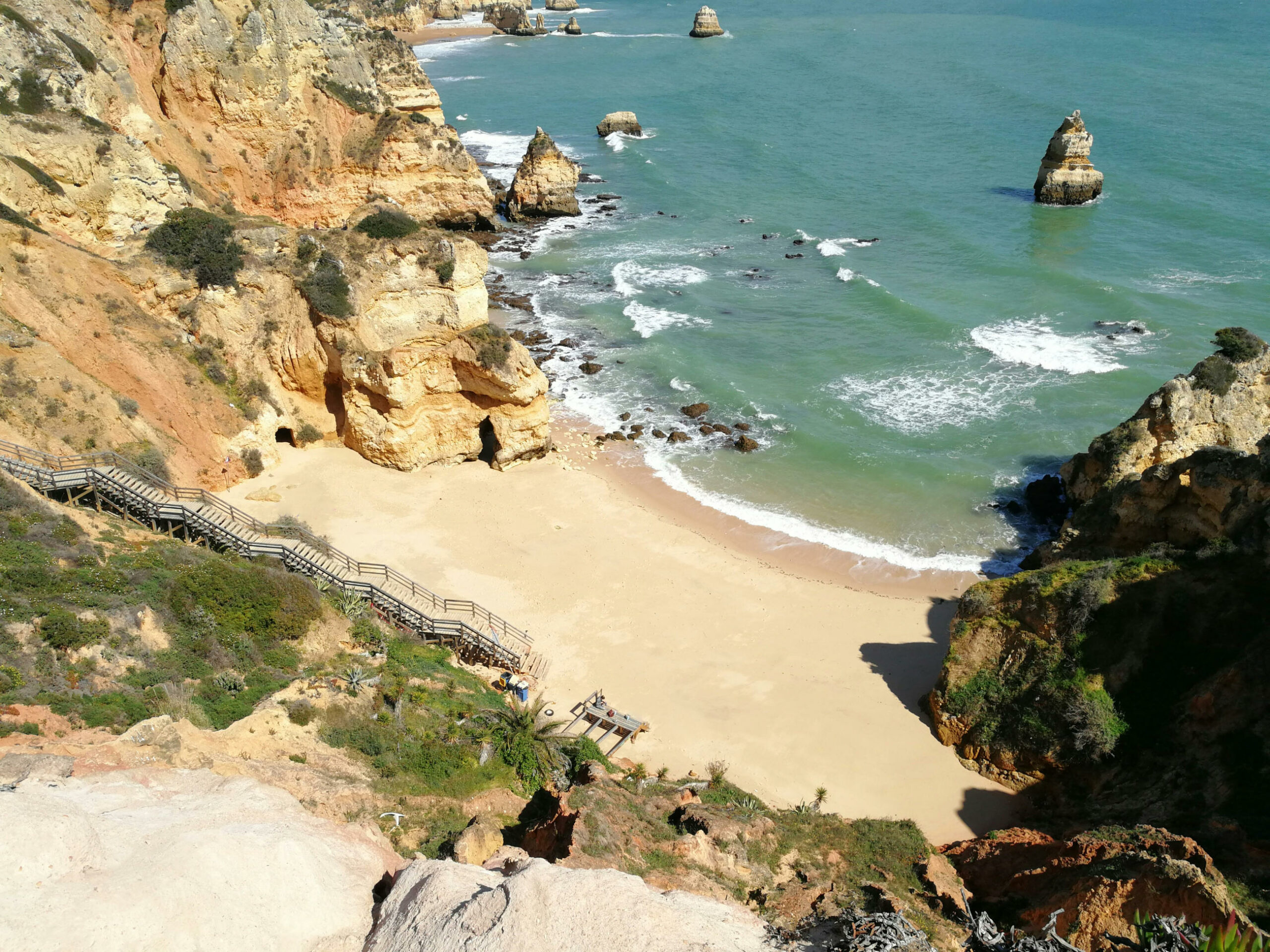 Das Bild zeigt die Praia do Camilo. Im Vordergrund ist ein begrünter Abhang mit einer Holztreppe. Mittig im Bild ist der Strand mit einigen Flesen. Im Hintergrund ist das Meer.